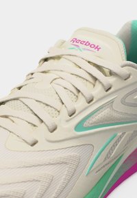 Κρέμα και πετρόλ αθλητικό παπούτσι Reebok με λευκά κορδόνια, επικαλύψεις από δίχτυ και συνθετικό υλικό, και ροζ λογότυπο στη γλώσσα.