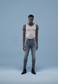 Világos bézs ujjatlan póló, szürke slim-fit farmernadrág, fekete bokacsizma; a modell egy egyszínű világos kék háttér előtt áll. Sima anyag, minimális textúrával.