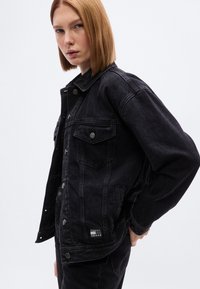 Μαύρο τζιν μπουφάν με χαλαρή εφαρμογή, με δύο τσέπες στο στήθος, μεταλλικά κουμπιά και λογότυπο στο κάτω αριστερό μέρος.