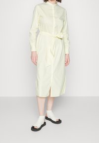 Vestito camicia lungo a maniche lunghe di colore giallo chiaro, con bottoni, cintura a fascia, spacco laterale e righe verticali. Abbinato a sandali bianchi con punta aperta.