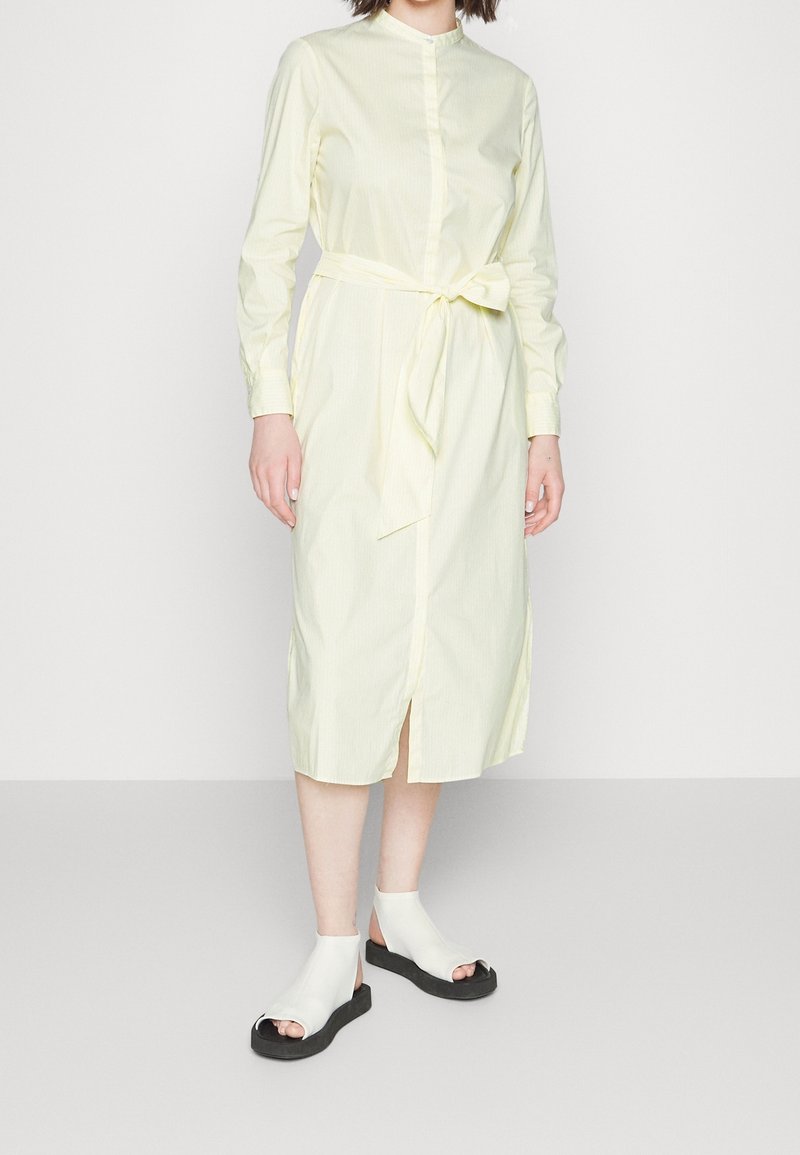 Vestito camicia lungo a maniche lunghe di colore giallo chiaro, con bottoni, cintura a fascia, spacco laterale e righe verticali. Abbinato a sandali bianchi con punta aperta.