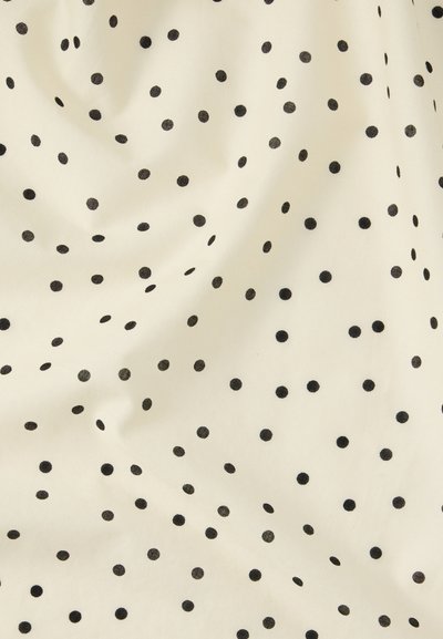 Ύφασμα κρεμ με μικρές μαύρες πουά διάσπαρτες, εμφανίζοντας ελαφριές πτυχώσεις και υφή.