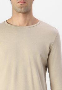 Camiseta de manga larga beige con cuello redondo y textura suave. Presenta un diseño ajustado con una apariencia lisa y minimalista.