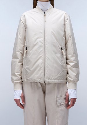 Osoba ubrana w beżową kurtkę typu bomber zapinaną na zamek z kieszeniami oraz beżowe spodnie cargo, ręce swobodnie opuszczone wzdłuż ciała na jasnym tle.