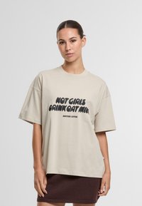 Camiseta de algodón beige con un corte holgado, mangas cortas y un texto negro audaz "NO A LAS CHICAS LES GUSTA LA LECHE DE AVENA" centrado en el frente.