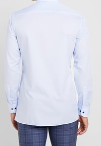 Světle modrá košile s dlouhými rukávy, hladkou texturou, zakřiveným lemem a kontrastními námořnickými knoflíky na manžetách. Jednoduchý pohled zezadu.