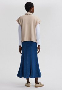 Beige gebreide top met cap sleeves, gelaagd over een wit shirt, gecombineerd met een lange blauwe spijkerrok. Het model draagt lichtgekleurde sneakers.
