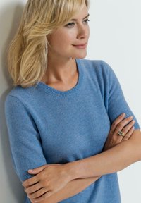 Blauer, kurzärmeliger Pullover aus weichem Material mit Rundhalsausschnitt und gerippten Bündchen, getragen von einer Person mit verschränkten Armen.