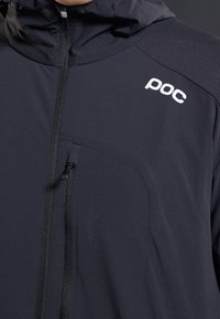 Giacca tecnica grigia scura con cappuccio, dotata di zip frontale, tasca laterale e logo bianco sul petto. Tessuto dalla texture liscia.