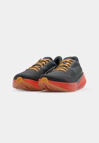 Chaussures de sport avec une tige en maille noire, des lacets orange et une semelle extérieure en caoutchouc rouge texturé, présentant un design épuré pour la performance.