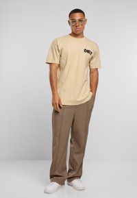 Beige bavlněné tričko s logem "OBEY" na levé části hrudi, v kombinaci s hnědými kalhotami a bílými teniskami. Nosí se čiré rámečkové brýle.