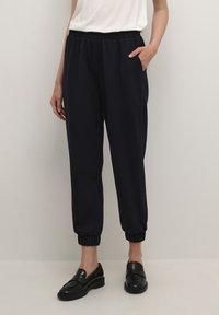 Persoon in zwarte joggerbroeken met elastische tailleband en gathered cuffs, met zwarte instappers, staand tegen een effen lichte achtergrond.