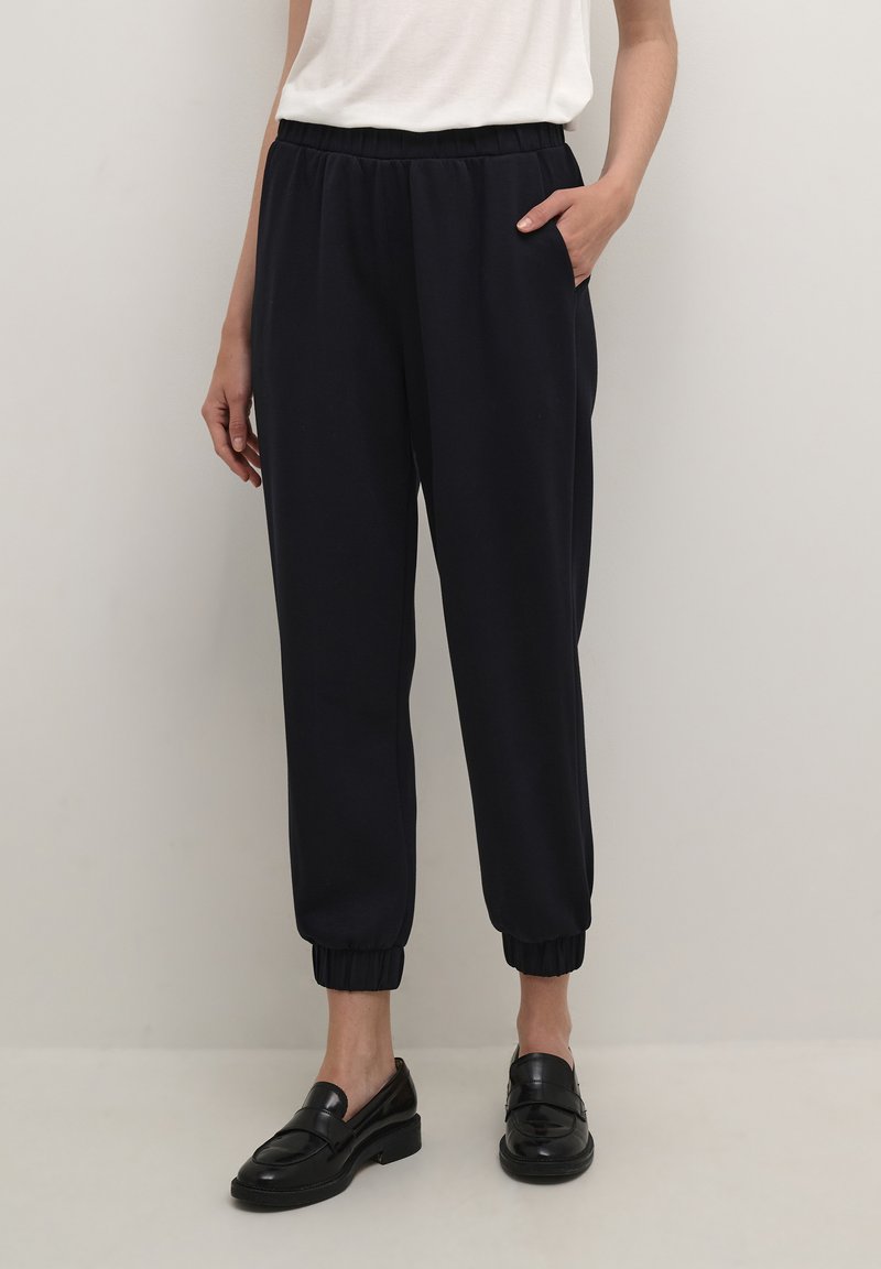 Persoon in zwarte joggerbroeken met elastische tailleband en gathered cuffs, met zwarte instappers, staand tegen een effen lichte achtergrond.