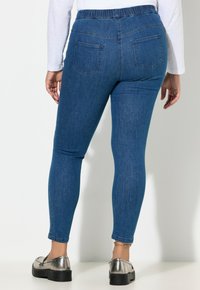 Niebieskie legginsy z denimu z elastycznym pasem, dwoma tylnymi kieszeniami i dopasowanym krojem, zestawione z metalicznymi butami na jasnym tle.