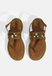 Braune Wildledersandalen mit einem T thong-Design, das drei runde, gehämmerte Goldakzente an den Riemen und eine strukturierte, braune Fußbettung aufweist.