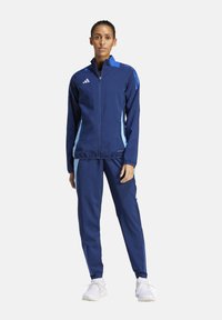 Marineblauwe trainingspak met lichtblauwe accenten, bestaande uit een jack met rits en bijpassende broek. Gemaakt van atletische stof met een gladde textuur.