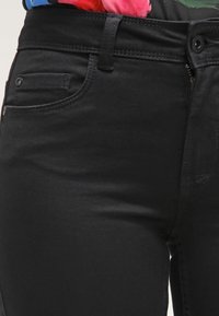 Jeans de mezclilla negra con un diseño ajustado. Presenta bolsillos frontales, presupuestos para cinturón y un cierre con cremallera. Tejido texturizado con un ligero brillo.