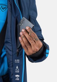 Marinejacke mit einem blauen Reißverschluss und Tags für wasserdichte und winddichte Eigenschaften; eine Hand, die ein schwarzes Smartphone in die Reißverschlusstasche steckt.