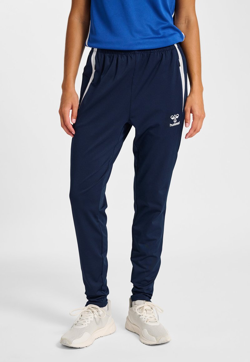 Persona che indossa pantaloni sportivi blu navy con strisce laterali bianche e logo, abbinati a scarpe da ginnastica bianche, in piedi su uno sfondo neutro.