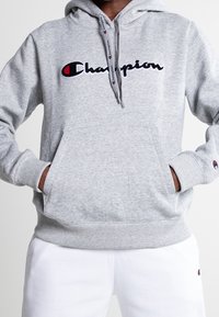 Sudadera de algodón gris con bolsillo frontal tipo canguro, puños y dobladillo acanalados, con el logo bordado de "Champion" en negro y rojo en el pecho.
