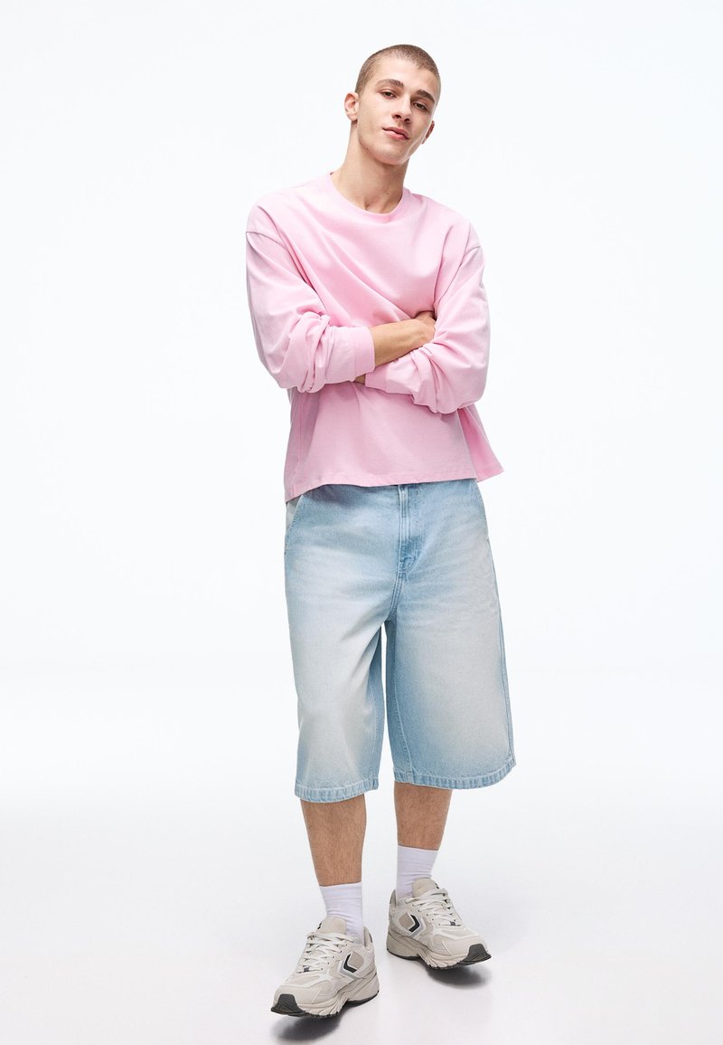 Giovane uomo con capelli corti che indossa una maglia rosa a maniche lunghe, pantaloncini di jeans azzurri, calzini bianchi e scarpe da ginnastica bianche, in piedi con le braccia incrociate.