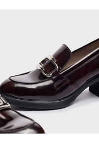 Mocasines de cuero burdeos con punta cuadrada, adornados con un detalle de hebilla plateada y un tacón bajo negro. Acabado suave y brillante.