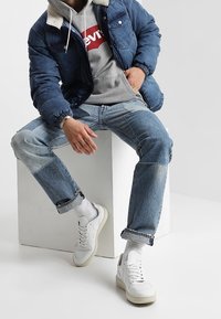 Chaqueta de denim con cuello de borreguillo, sudadera gris con logo rojo, jeans de patchwork y zapatillas blancas. Un conjunto casual y en capas.
