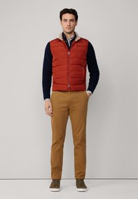 Chaleco acolchado rojo con cierre de cremallera sobre un suéter azul marino, combinado con pantalones beige y zapatillas olivas. Tela suave y diseño casual.