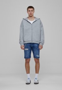 Pelēka zip-up kapuce ar lielu kapuci, kopā ar nodilušām zilu džinsu šortiem. Valkāta ar baltām zeķēm un melni baltām kedām.