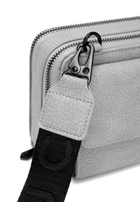 Detailní záběr na roh světle šedé texturované tašky s černým kovovým uzávěrem, který připojuje černý látkový popruh k táhlu zipu.