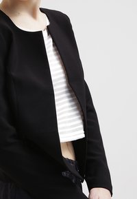 Veste noire courte avec un col rond et une ouverture devant, superposée à un crop top rayé blanc et gris. Texture de tissu lisse.