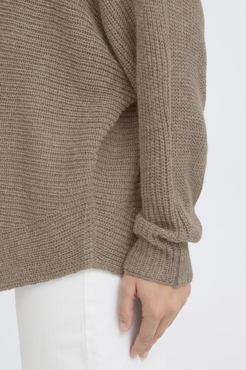 Gros plan d'une main reposant à côté d'une manche de pull beige en tricot et d'un pantalon blanc sur un fond uni.