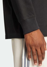 Black long-sleeve shirt with a ribbed cuff, displaying a textured material. A hand rests on light-colored pants featuring navy stripes.