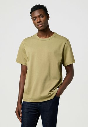 Oliven grøn bomulds t-shirt med korte ærmer, rund halsudskæring og afslappet pasform. Bæret med mørke denim jeans, hånden i lommen. Enkel design.