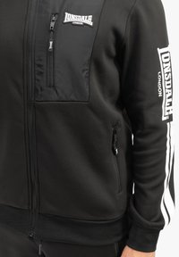 Melnā zipa jaka ar teksturētu melnu kabatu, baltiem svītrojumiem uz piedurknēm un Lonsdale logo detaļām uz krūtīm un piedurknēm.