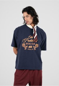 Tamsiai mėlyna trumpo rankovio marškinėliai su auksinės spalvos grafika, vaizduojančia liūtus ir užrašu "Draudžiamas socialinis klubas Berlyne". Pagaminta iš medvilnės, laisvas kirpimas.