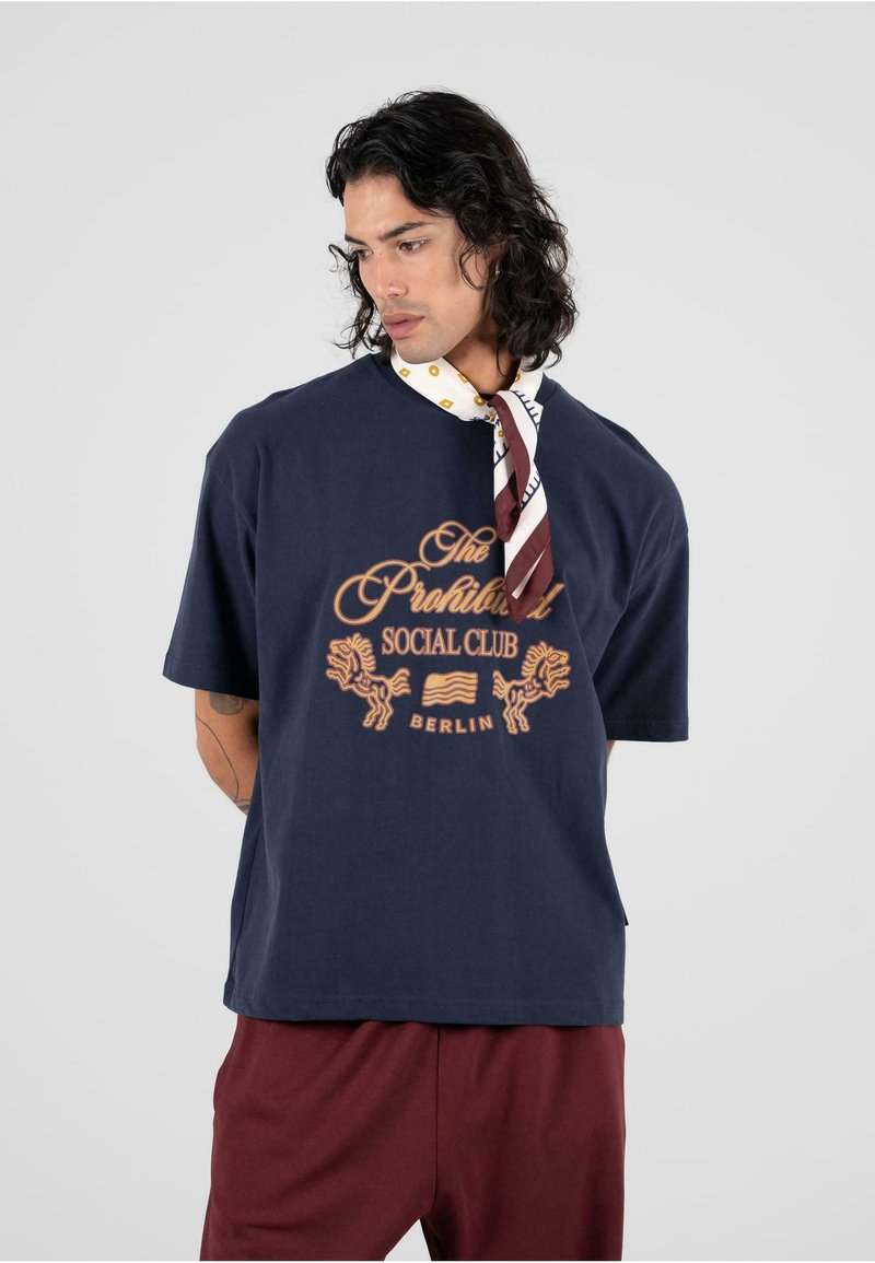 Tamsiai mėlyna trumpo rankovio marškinėliai su auksinės spalvos grafika, vaizduojančia liūtus ir užrašu "Draudžiamas socialinis klubas Berlyne". Pagaminta iš medvilnės, laisvas kirpimas.