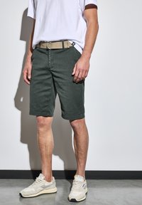 Pantalones cortos de color verde oscuro con un corte estándar, que cuentan con una cremallera y bolsillos laterales, combinados con un cinturón beige y zapatillas de color claro.