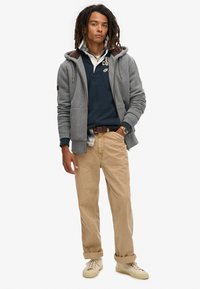 Grijze rits hoodie met een fleece-voering in de capuchon, gedragen over een marineblauwe polo, kaki broek en beige sneakers. Opvallende details zijn een geruite shirt dat eronder valt.