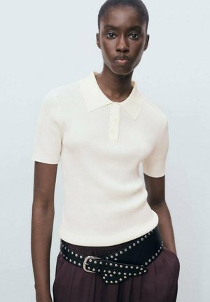Crème polo-shirt met korte mouwen en ribbeltextuur, voorzien van een puntkraag. Gestyled met een zwarte studsriem over donkere broeken.