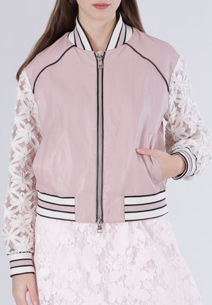 Femme portant une veste bombardier rose pâle avec des manches en dentelle florale transparente et un col, des poignets et un ourlet côtelés rayés, main dans la poche.