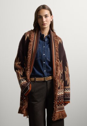 Giovane donna che indossa un cardigan marrone con frange e motivo sopra una camicia blu con bottoni e pantaloni scuri con cintura beige.