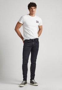 Muškarac nosi bijelu majicu Pepe Jeans, crne uske traperice i sive tenisice, a stoji ispred jednostavne svijetlosive pozadine.