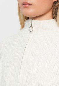 Bijela pletena sweatertas s teksturiranim uzorkom pletenice, visokim ovratnikom i metalnim zatvaračem s okruglim povlačnim gumbom.
