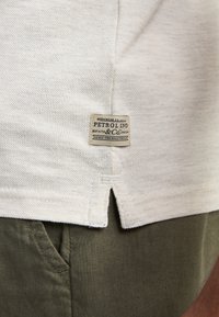 Ourlet de chemise en tissu beige texturé avec une étiquette de la marque Petrol Ind, associé à un pantalon vert olive montrant la couture de la poche et la texture du tissu.