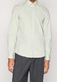 Lichtgroene blouse met lange mouwen, een subtiel verticaal patroon, knoopsluiting en een puntige kraag. Draag met grijze ribbelbroeken.