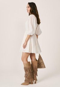 Mujer vestida con un vestido blanco de mangas abullonadas, botas beige con flecos hasta la rodilla, y sosteniendo un bolso marrón con estampado, de pie contra un fondo liso.