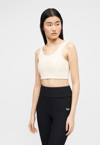 Νεαρή γυναίκα με μακριά σκοτεινά μαλλιά που φοράει ένα μπεζ κοντό τοπ και μαύρο κολάν με ψηλή μέση και λογότυπο στη μέση.