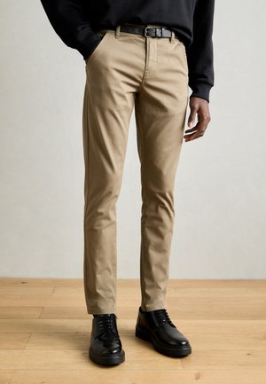 Homme portant un pantalon beige slim, ceinture noire, haut noir à manches longues et chaussures habillées noires brillantes sur un sol en bois.