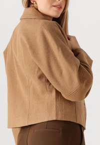 Jas van kamelenwolmix met een ontspannen pasvorm, voorzien van een reverskraag en een gestructureerd oppervlak, gecombineerd met bruine, op maat gemaakte broeken.